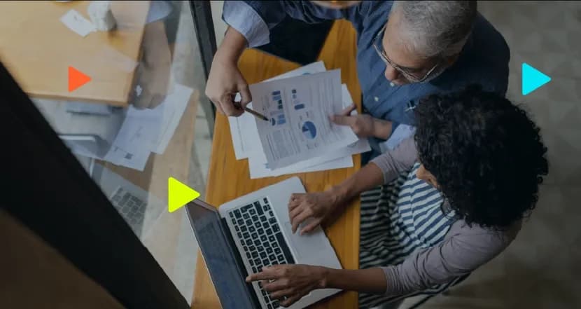 Visão de cima de um homem e uma mulher analisando gráficos impressos e usando um notebook sobre uma mesa de madeira. Ícones coloridos de 'play' aparecem sobrepostos à imagem
