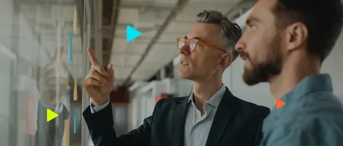 Dois profissionais analisando painel de vidro com post-its, representando produtividade e controle com o Ploomes.