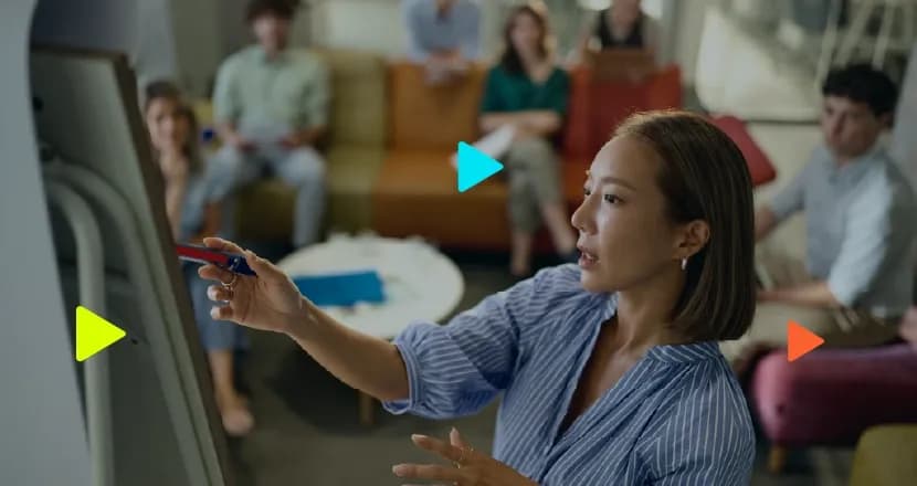 Mulher asiática de camisa azul listrada faz apresentação em pé, apontando com caneta para um quadro. Ao fundo desfocado, pessoas sentadas em sofás assistem à reunião num escritório descontraído.