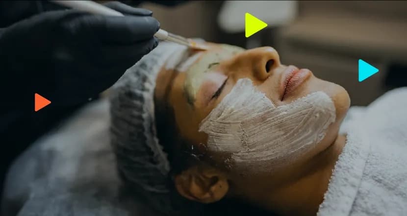 Profissional com luvas pretas aplicando máscara facial com pincel em cliente deitada em clínica de estética.
