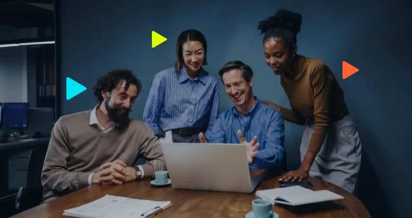 Quatro profissionais sorrindo enquanto olham juntos para a tela de um laptop em uma mesa redonda de reunião.
