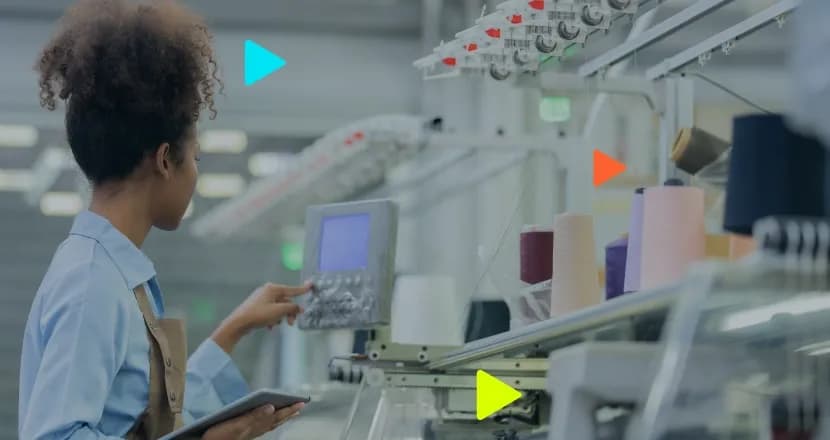 Mulher negra com cabelo preso e avental marrom segura um tablet enquanto opera o painel digital de uma máquina têxtil na fábrica. Ao fundo, veem-se carretéis de linha coloridos.