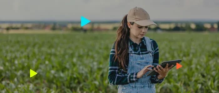 Mulher de boné e jardineira jeans usa tablet em meio a uma plantação verde.