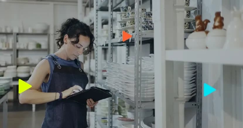 Mulher de avental usa tablet para checar estoque em prateleiras de cerâmica.