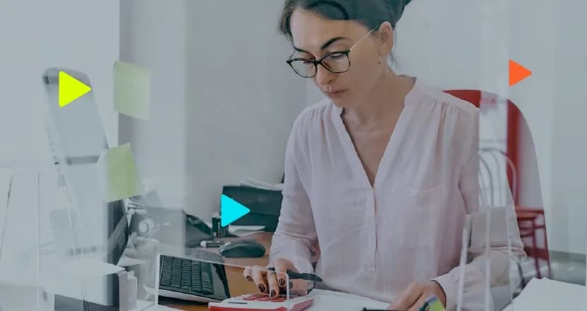 Mulher com óculos e camisa rosa claro sentada a uma mesa de escritório, usando uma calculadora rosa. Ao lado, um monitor de computador com post-its e formas geométricas coloridas (ícones de 'play') sobrepostas à imagem. Foco está na ação da calculadora.