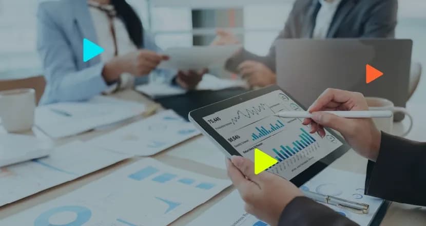 Mãos de uma pessoa segurando um tablet que exibe gráficos financeiros de barras e linhas com indicadores numéricos, apontando para a tela com uma caneta stylus branca. Ao fundo, desfocado, profissionais em trajes formais participam de uma reunião ao redor de uma mesa com relatórios impressos. Ícones geométricos coloridos em formato de 'play' aparecem sobrepostos na imagem.