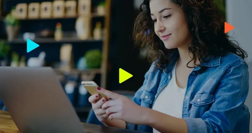 Mulher jovem de cabelos cacheados escuros, vestindo jaqueta jeans e blusa branca, sorrindo enquanto mexe em um smartphone. Ela está sentada de frente para um notebook aberto sobre uma mesa de madeira. Ao fundo, desfocado, há uma estante com objetos e plantas. Ícones geométricos coloridos em formato de 'play' aparecem sobrepostos à imagem.