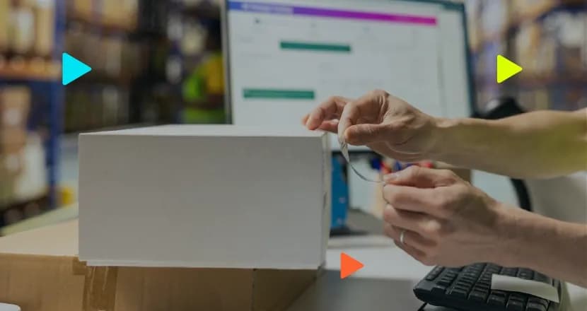 Mãos colando uma etiqueta em uma caixa branca. Ao fundo, um monitor de computador e prateleiras de estoque desfocadas, com ícones coloridos de 'play' na imagem.