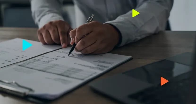 Close das mãos de um profissional com camisa de manga comprida assinando um documento de papel com caneta sobre uma prancheta em uma mesa de madeira, com um notebook fechado ao lado.