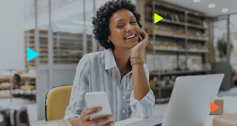 ulher negra sorridente segurando um celular em frente a um notebook em um escritório. Ícones coloridos de 'play' sobrepostos à imagem.