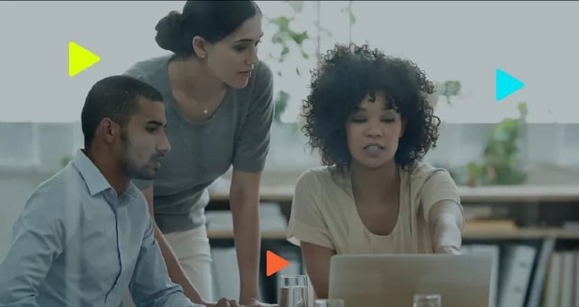 Três profissionais reunidos analisando a tela de um notebook em um escritório. Ícones coloridos de 'play' aparecem sobrepostos à imagem.