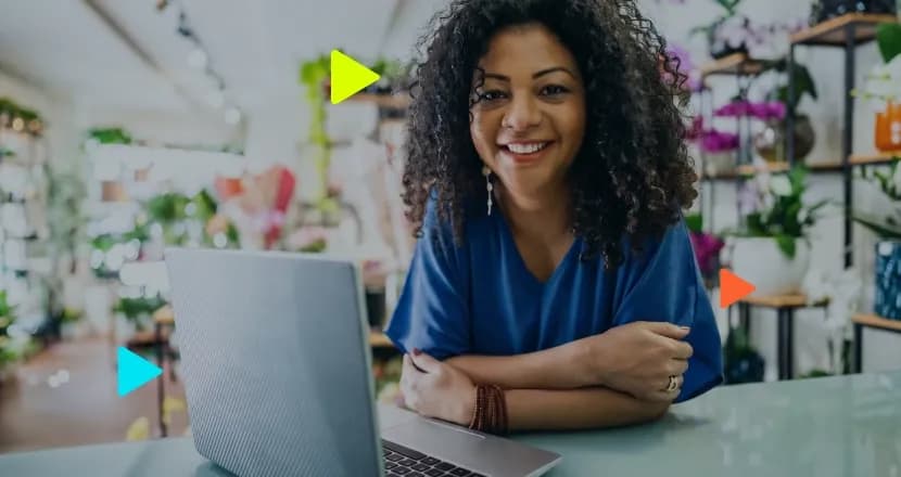 Mulher sorridente de cabelos cacheados sentada com um notebook em uma floricultura. Ícones coloridos de 'play' sobrepostos à imagem
