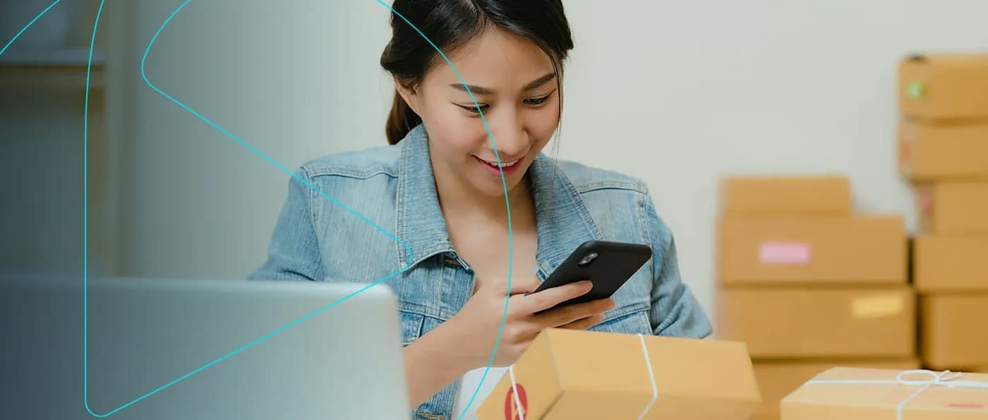 Vendedora conferindo um pedido no celular entre caixas de envio e o notebook