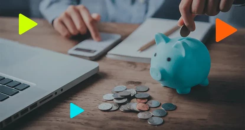 Pessoa colocando moeda em cofrinho de porquinho azul sobre mesa com moedas espalhadas, calculadora e notebook ao fundo.