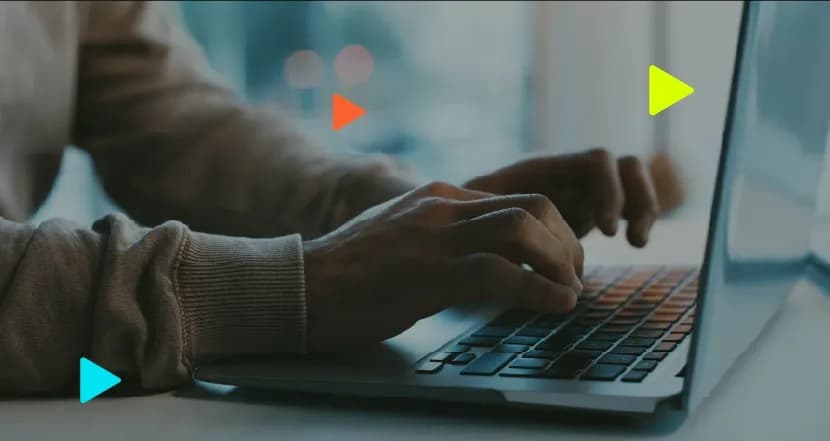 Close em mãos de uma pessoa vestindo blusa de manga longa digitando no teclado de um notebook. Ícones coloridos de 'play' aparecem sobrepostos à imagem com fundo desfocado.