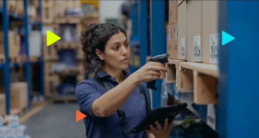 Mulher vestindo uniforme azul trabalhando em um estoque, segurando um tablet e usando um leitor de código de barras para escanear etiquetas em caixas de papelão nas prateleiras. Ícones coloridos de 'play' aparecem sobrepostos à imagem.