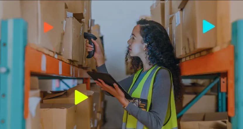Mulher de cabelos cacheados vestindo colete refletivo trabalhando em um estoque. Ela segura um tablet e usa um leitor de código de barras para escanear uma caixa de papelão armazenada em uma prateleira. Ícones coloridos de 'play' aparecem sobrepostos à imagem.