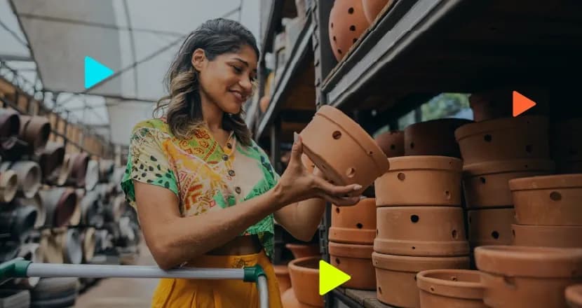 Mulher sorridente vestindo uma blusa estampada colorida e calça amarela, segurando e observando um vaso de cerâmica em uma loja com prateleiras repletas de itens semelhantes. Ela está encostada em um carrinho de compras. Ícones coloridos de 'play' aparecem sobrepostos à imagem.