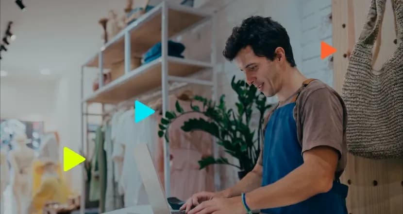 Homem sorridente vestindo avental azul digitando em um notebook no balcão de uma loja de roupas. Ao fundo, araras com peças de vestuário, uma planta e uma bolsa pendurada na parede. Ícones coloridos de 'play' aparecem sobrepostos à imagem.