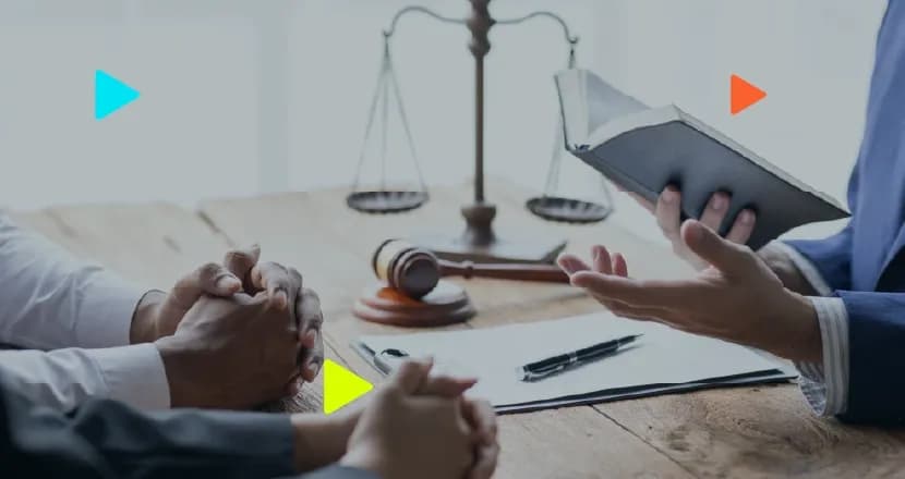Advogado segurando livro durante reunião com clientes em mesa com martelo e balança da justiça.