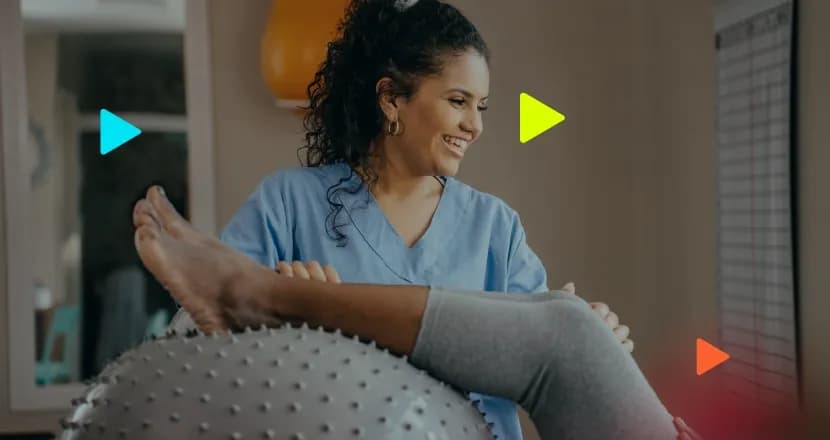 Fisioterapeuta sorridente auxiliando paciente em exercícios de reabilitação com bola de equilíbrio.