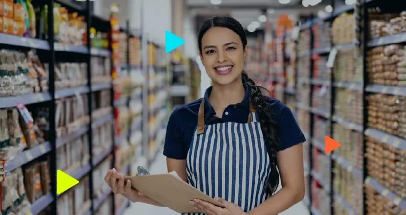 Mulher jovem e sorridente, de avental listrado e camisa azul, segura uma prancheta em um corredor de supermercado. Ao fundo desfocado, prateleiras repletas de produtos de mercearia.