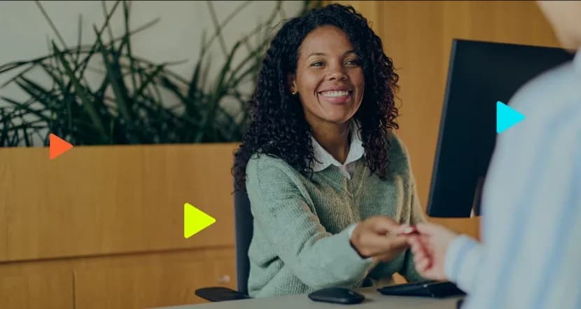 Recepcionista sorridente entregando objeto para cliente em balcão de atendimento com computador.