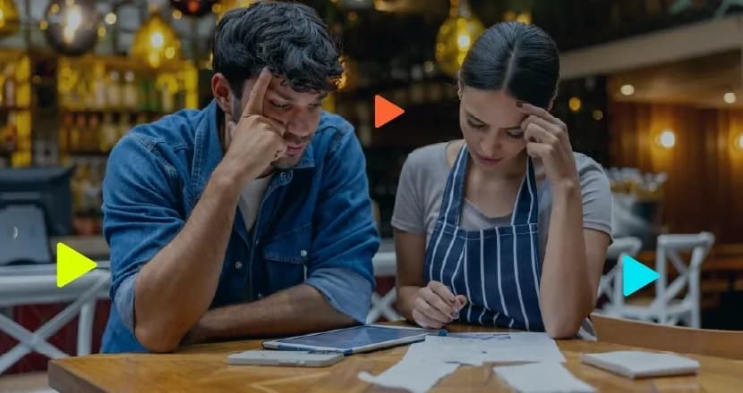 Homem e mulher de avental sentados à mesa de um restaurante, olhando preocupados para papéis e contas sobre a mesa.