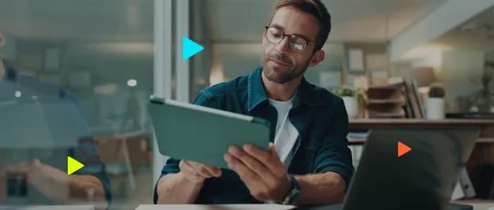 Homem de óculos usa tablet verde em escritório moderno iluminado. Um notebook aparece em primeiro plano à direita.