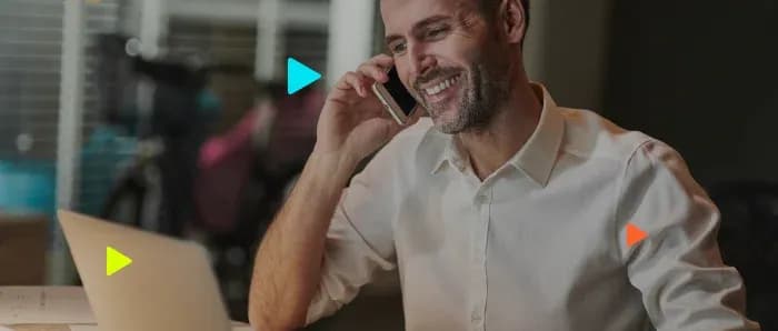 Homem sorrindo ao falar no celular em frente a um notebook aberto em ambiente de escritório corporativo.
