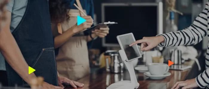 Cliente aponta para tela de tablet no balcão de uma cafeteria com atendentes ao fundo.