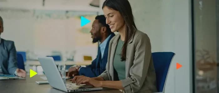 Mulher sorrindo enquanto digita no notebook em uma mesa de reunião com colegas ao fundo.