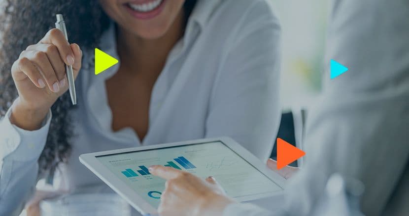 Mulher sorridente com uma caneta na mão analisando gráficos financeiros na tela de um tablet junto com outra pessoa que aponta para o dispositivo.