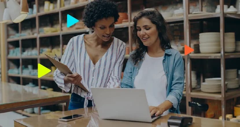 Duas mulheres conversam diante de um laptop em um balcão de atendimento, com prateleiras de produtos ao fundo.