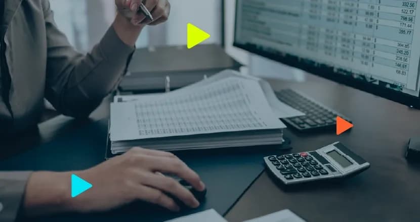 Mãos de um profissional trabalhando em mesa de escritório utilizando mouse e segurando caneta, com pilha de documentos impressos, calculadora e monitor exibindo planilha.