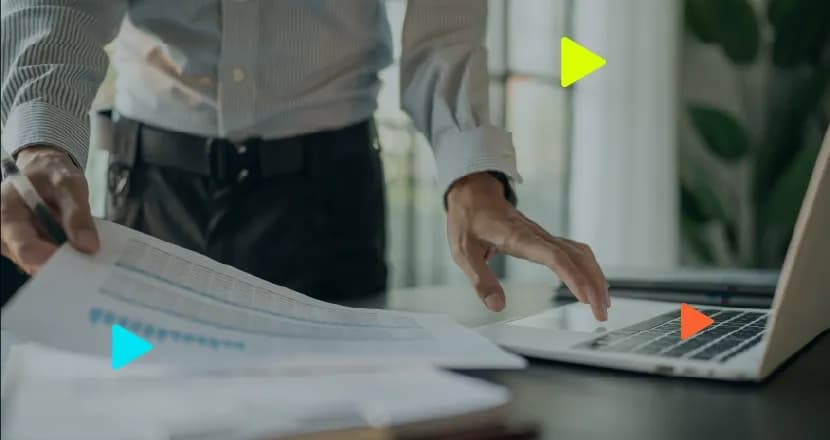 Profissional analisa documento impresso com tabela de dados em uma mão enquanto utiliza um notebook sobre a mesa de trabalho.