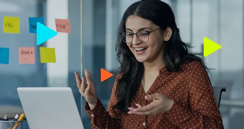 Mulher sorrindo faz videochamada no notebook em escritório com notas adesivas coloridas ao fundo.