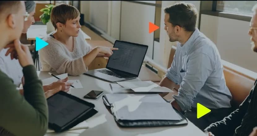 Mulher aponta para tela de notebook durante reunião de negócios com colegas ao redor de uma mesa com papéis.