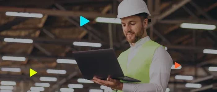 Engenheiro de capacete e colete usando notebook dentro de um grande galpão industrial.