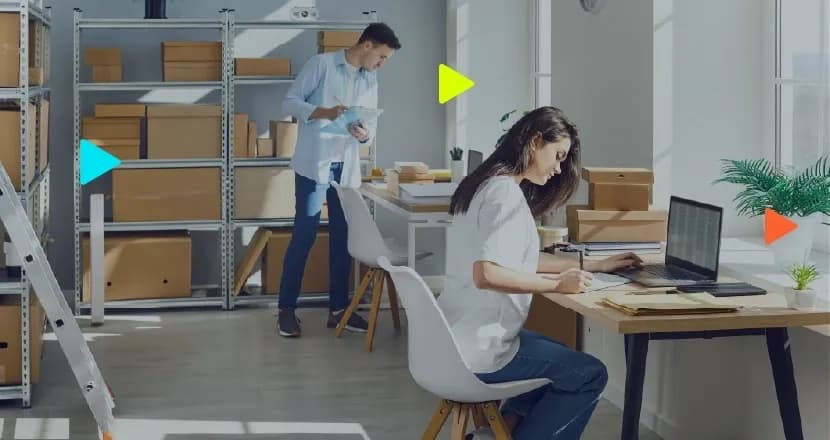 Mulher usa notebook em mesa e homem checa caixas em prateleira de metal com prancheta, em ambiente de logística.