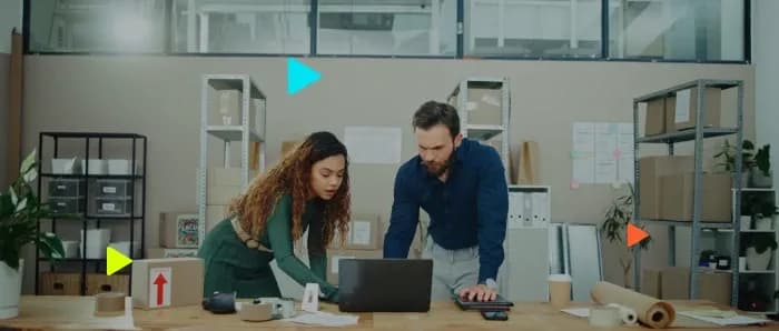 Homem e mulher gerenciam logística de e-commerce no notebook. Caixas de papelão e prateleiras ao fundo do escritório.