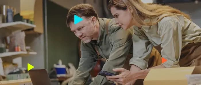 Homem e mulher de avental olham para tela de notebook em oficina, com caixas de papelão ao lado.