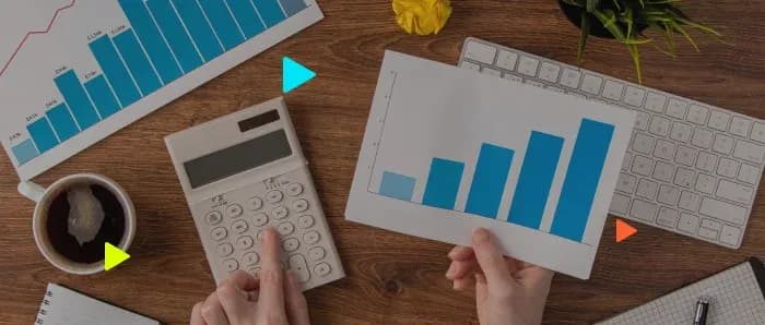 Vista superior de uma pessoa trabalhando em uma mesa de madeira. Ela usa uma calculadora com uma mão e segura um gráfico de crescimento com a outra, cercada por itens de escritório como teclado, café e papéis.