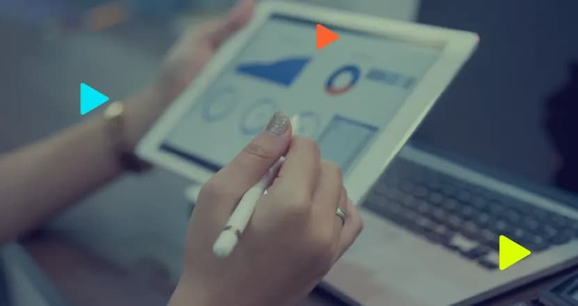 Mãos de profissional usam caneta digital em tablet exibindo painel de gráficos financeiros, com notebook desfocado ao fundo.
