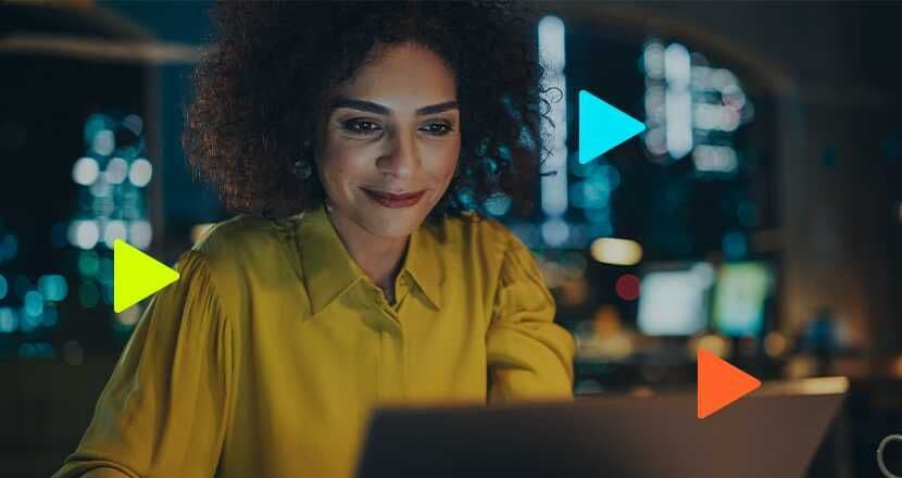 Mulher com cabelo cacheado, usando blusa amarela, sorri enquanto trabalha no laptop em um escritório à noite.