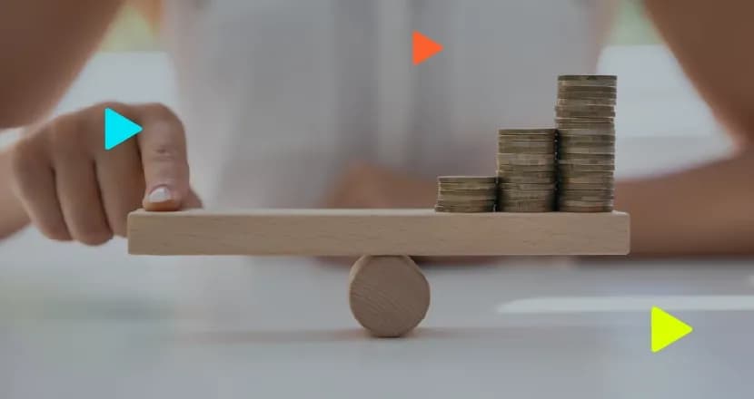Dedo equilibra tábua de madeira em formato de gangorra com pilhas de moedas, ilustrando o ponto de nivelamento financeiro.
