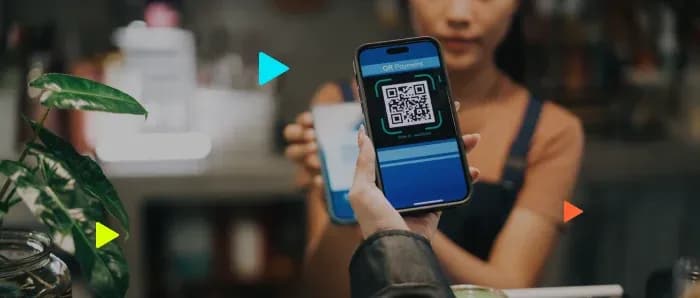 Mão segurando celular com QR Code de pagamento na tela em frente ao caixa de um estabelecimento.