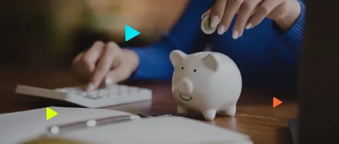 Pessoa coloca moeda em cofrinho de porco branco enquanto usa uma calculadora na mesa.