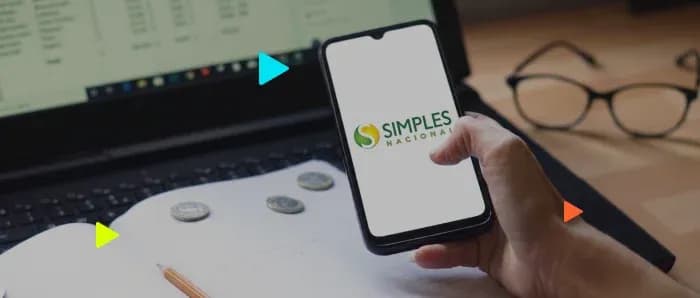 Mão segura celular exibindo a logo do Simples Nacional. Ao fundo, notebook, moedas sobre caderno e óculos de grau.