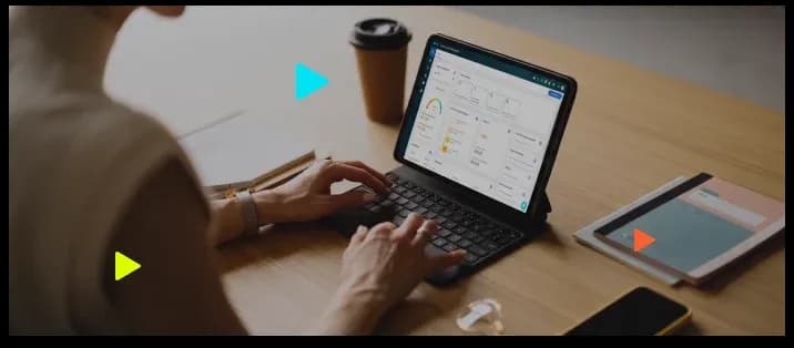 Mulher digita em tablet com teclado em mesa de madeira com café e caderno.